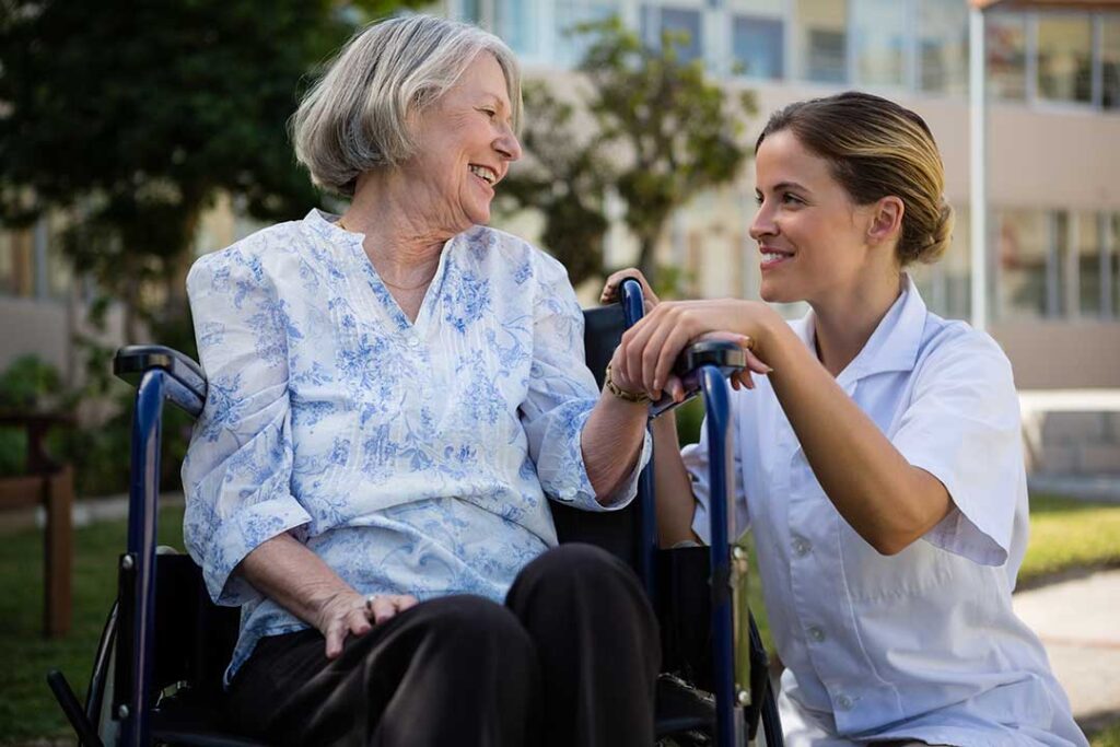 An elderly woman and caregiver smiling together in a comfortable, tidy common area.